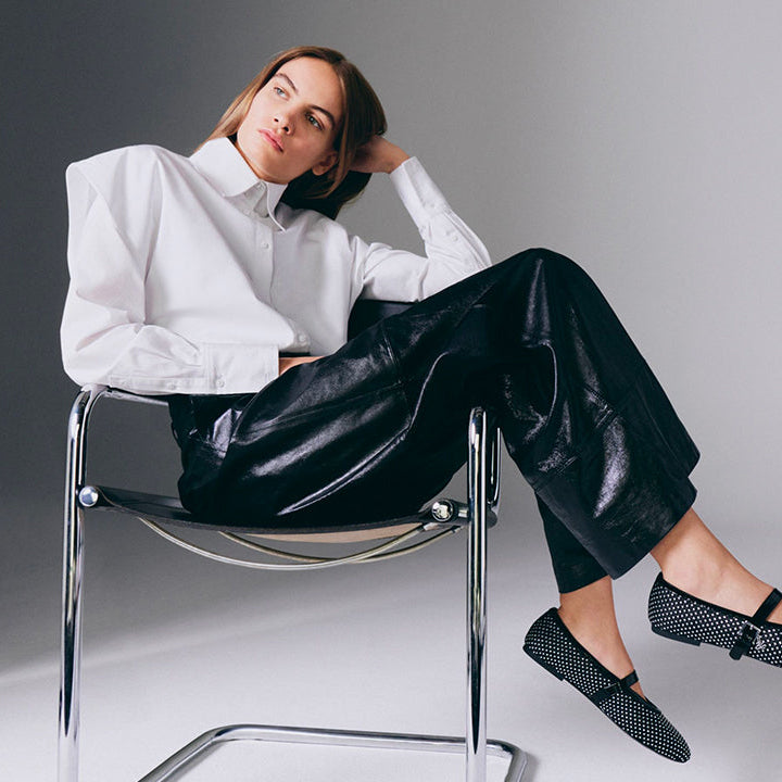 Karl Lagerfeld campaign: a model seated in a chair wearing a white shirt and black leather trousers with studded flats.